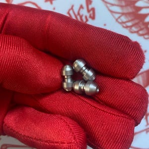 May include: Three small, silver-colored metal objects with a cylindrical shape and rounded ends. The objects are held in a red fabric. The objects appear to be components or parts of a larger item.