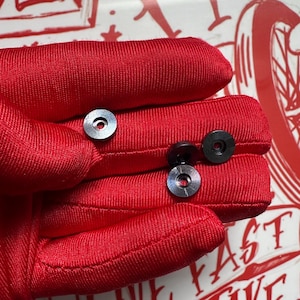 Puede incluir: Primer plano de varias arandelas met&aacute;licas negras peque&ntilde;as con un agujero central y un anillo exterior plateado. Las arandelas est&aacute;n sobre una superficie de tela roja. La imagen est&aacute; bien iluminada, resaltando los detalles.