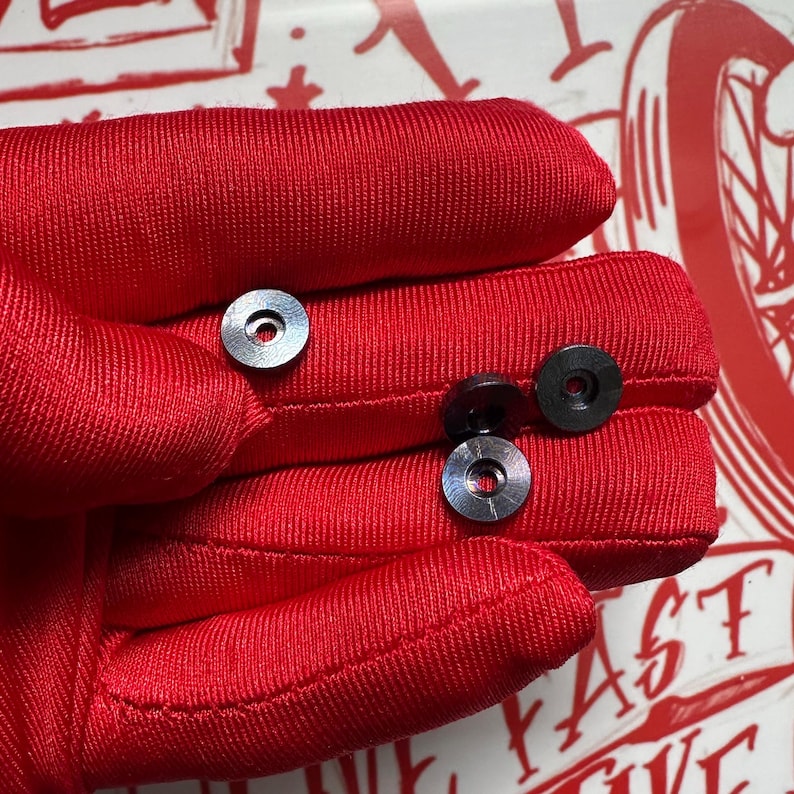 Puede incluir: Primer plano de varias arandelas met&aacute;licas negras peque&ntilde;as con un agujero central y un anillo exterior plateado. Las arandelas est&aacute;n sobre una superficie de tela roja. La imagen est&aacute; bien iluminada, resaltando los detalles.
