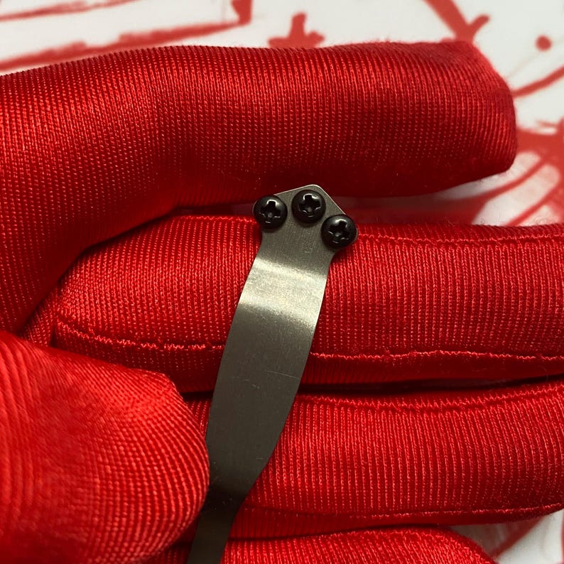 Puede incluir: Primer plano de un clip de navaja de metal plateado con tres tornillos negros. El clip se encuentra sobre un fondo de tela roja vibrante, creando un contraste llamativo. El clip de la navaja es un peque&ntilde;o accesorio funcional.