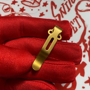 May include: A gold-colored pocket clip with a rectangular shape and a curved end. The clip has a central elongated opening and two circular holes at the top. The clip is held against a red fabric background.