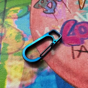 May include: A blue and black carabiner clip. The clip has a curved design with a straight bar and a rectangular opening. The background is a colorful fabric with graffiti-style art.