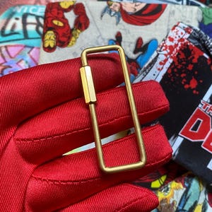 May include: A rectangular brass key holder with a screw-in post. The key holder is held against a background of red fabric and comic book fabric.