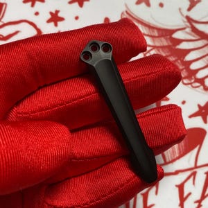 May include: A black metal object with three circular holes at one end and a long, slender body. The object is held against a red fabric background, creating a contrast of textures and colors. The design suggests a tool or component.