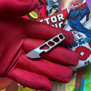 May include: A small, matte grey metal utility blade tool featuring a skeletonized handle with three distinct cutouts and a pointed tip, resting against a bright red fabric surface with a comic book graphic print.