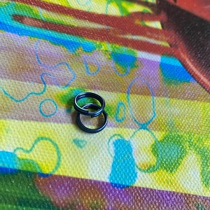 May include: Two small, dark blue metal split rings are interlocked, resting on a colorful, patterned surface. The rings are circular and appear to be made of a solid, durable material. The background features a mix of colors, including yellow, blue, red, and purple.