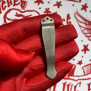 May include: A metallic pocket knife clip featuring an engraved koi fish pattern. The clip has a tapered shape with three circular mounting holes at the top and a pointed tip at the bottom, showcasing a matte grey finish with detailed line art.