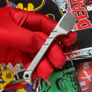 May include: A matte gray metal pry bar tool featuring a flat, angled chisel tip, a textured handle with diagonal grooves, and a circular lanyard hole at the base. The tool has a sleek, industrial design with a uniform finish and a sturdy construction.