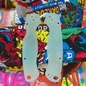 May include: Two translucent green knife handle scales with multiple holes and colored accents. The scales are set against a colorful background of comic book fabric featuring Marvel characters and text.