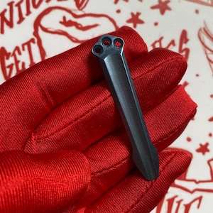 May include: A dark gray metal object with three circular holes at one end and a pointed tip. The object is held in a red glove, with a design visible in the background.