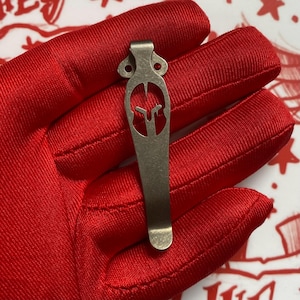 Puede incluir: Un clip de bolsillo de color bronce con un dise&ntilde;o de casco espartano. El clip se sostiene sobre un fondo de tela roja. El clip tiene una forma curvada con una parte superior decorativa y un dise&ntilde;o ranurado.