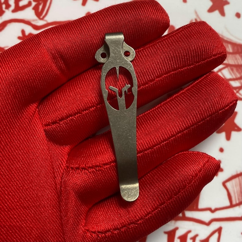 Puede incluir: Un clip de bolsillo de color bronce con un dise&ntilde;o de casco espartano. El clip se sostiene sobre un fondo de tela roja. El clip tiene una forma curvada con una parte superior decorativa y un dise&ntilde;o ranurado.
