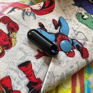 May include: A black, cylindrical object with a hole, next to a light blue button. The objects are on a fabric with a comic book print featuring characters like Spider-Man, Thor, and Iron Man.