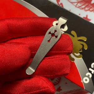 May include: A silver-colored metal clip with a decorative cross cutout. The clip is held against a red fabric background. The clip has a curved top and a rounded bottom. The background also includes a black and red card with gold accents.