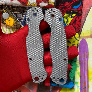 May include: Two silver-colored, textured knife scales with a diamond pattern. The scales have several holes and are set against a backdrop of red fabric and colorful comic book-style illustrations. The scales are designed for a knife.