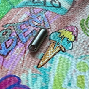 May include: A small, silver metal capsule with a circular opening at one end. The capsule is positioned on a colorful fabric with graffiti-style lettering and a cartoon ice cream cone illustration. The fabric has a mix of pink, blue, green, and yellow colors.