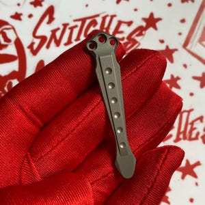 May include: A small, gray metal object with a series of evenly spaced, circular indentations down its length. The top portion features three circular holes. The object is held against a red background with white text and star shapes.