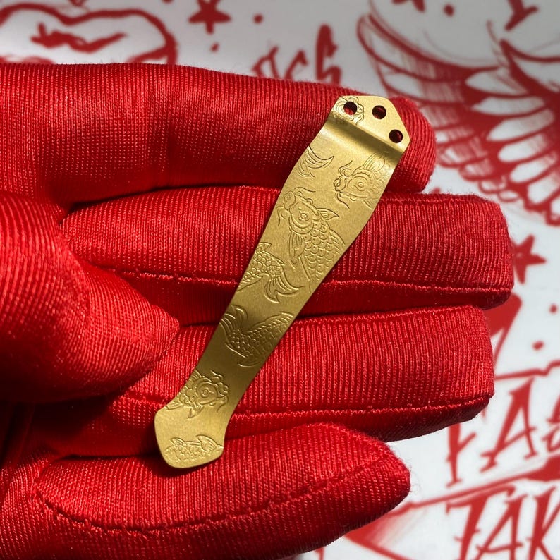 May include: A gold-toned metal pocket clip featuring an engraved koi fish pattern. The elongated, slightly curved clip has three small circular mounting holes at the top end and a rounded base, showcasing detailed scale and fin textures throughout.