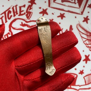May include: A metallic bronze-toned pocket knife clip featuring an intricate laser-engraved pattern of multiple koi fish swimming in various directions. The clip has a tapered shape with two small circular mounting holes at the top end.