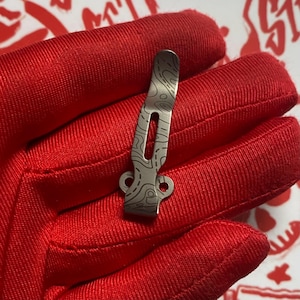 May include: A metallic pocket clip featuring a dark gray finish and a laser-etched topographic map pattern. The clip has a slender, elongated body with a central cutout and a wider base section containing two circular mounting holes for attachment.