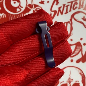 May include: A close-up of a dark purple metal clip. The clip has a curved top with two small circular holes and a long, rectangular body with a vertical slot. The clip is held against a red fabric background.