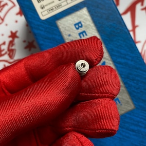 May include: A small, silver-colored knife pivot screw held in a red gloved hand. The screw has a circular head with concentric rings. A blue box with the word "BE" is in the background.