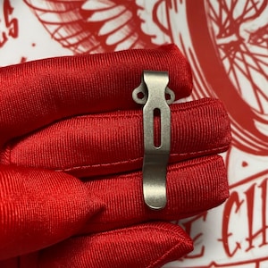 May include: A silver-colored metal pocket clip with a rectangular slot and two circular holes. The clip is held against a red fabric background.
