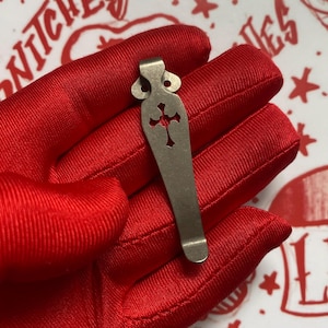 May include: A silver-colored metal clip with a cross-shaped cutout. The clip has a decorative top with two circular cutouts. The clip is held in a red glove.