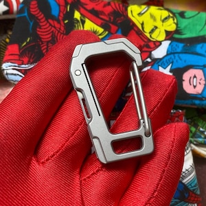 May include: A silver-colored, rectangular carabiner clip with a spring-loaded gate. The clip has a built-in tool and a sturdy design. The background is a red surface with a colorful comic book print.