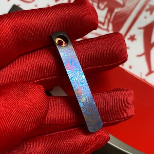 May include: A rectangular metal pendant with a swirling pattern of blue, purple, and gold hues. The pendant has a small hole at one end and is displayed against a red fabric background.