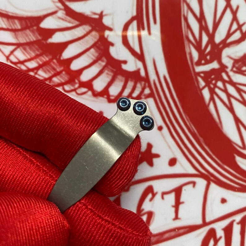 May include: A close-up of a silver-colored metal clip with three small, dark blue screws. The clip is held against a red fabric background. The clip appears to be a part of a larger object.