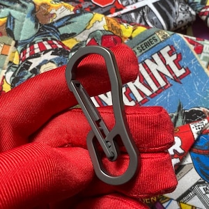 May include: A dark gray metal carabiner with a smooth, elongated oval shape and a spring-loaded gate mechanism. The carabiner is held against a background featuring a colorful comic book print with red, yellow, and blue graphic elements.