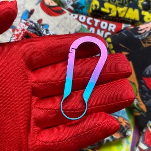 May include: A small, colorful carabiner clip with a gradient finish, transitioning from purple to blue. The clip is held against a red fabric background, with a comic book print visible in the background.