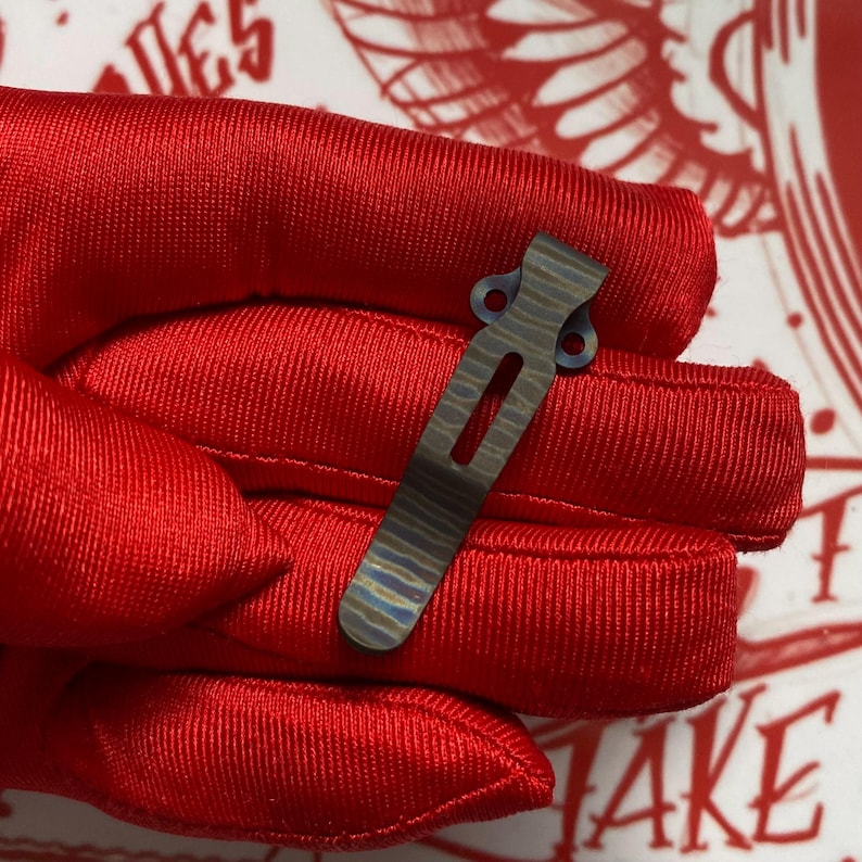 May include: A close-up of a small, metallic pocket clip with a striped pattern in shades of blue, gray, and gold. The clip has a rectangular shape with a long, narrow opening and two small holes at the top. The clip is held against a red background.