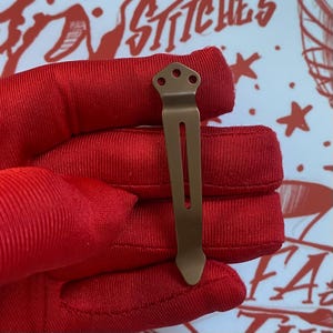 May include: A matte bronze-colored metal pocket clip featuring a pointed tip and a flat top section with three circular screw holes. The clip has a long, narrow vertical slot running through the center of its body, designed for attachment to gear.
