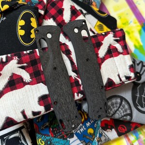 May include: Two textured black knife scales with multiple holes and cutouts. The scales are set against a colorful background of patterned fabric, including a Batman logo and a red and black plaid design with white bear silhouettes.