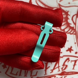 May include: A close-up shot of a light teal pocket clip. The clip is made of metal and has a rectangular shape with a curved top and a slot down the center. It has two small holes near the top. The clip is held against a red fabric.