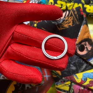 May include: A silver-colored key ring is held against a red fabric background. The key ring is circular with a small opening. The red fabric has a textured appearance. The background features a colorful pattern.