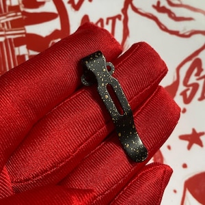 May include: A black metal pocket clip with gold speckles. The clip has a curved shape with a long, narrow opening in the center and a small loop at the top. The clip is shown against a red fabric background.