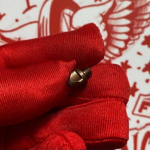 May include: Close-up of a small, bronze-colored screw with a hexagonal head and threaded shaft. The screw is held against a vibrant red fabric, creating a contrast in textures and colors. The background is a blurred white and red pattern.
