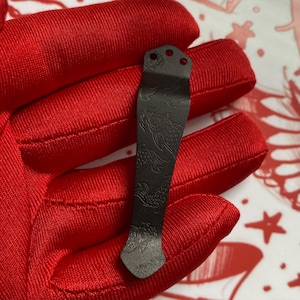 May include: A dark gray metal pocket clip with a decorative fish scale pattern. The clip has three holes at the top and a curved design. The clip is held in a red glove.