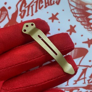 May include: A matte gold-toned metal pocket clip featuring a rounded top with three small circular holes and a long, narrow vertical slot running down the center of the body, ending in a tapered, slightly pointed tip for secure attachment.