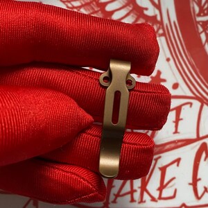 May include: A bronze-colored pocket clip with a rectangular slot and two circular holes. The clip is held against a red fabric background. The clip is likely for a folding knife or similar item.
