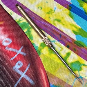 May include: A silver and gold-toned metal tool with a sharp, pointed end. The tool has a textured grip and a small hole at the top. The background is a colorful abstract design with red, blue, yellow, and purple hues.