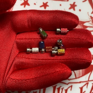 May include: A collection of small, colorful metal objects with screw-like ends. The objects are in various colors, including red, green, blue, and gold. They are displayed on a red surface.