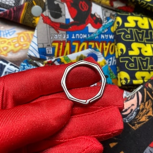 May include: A silver-colored, geometric ring with an octagonal design. The ring is held against a vibrant red fabric, with a colorful comic book-themed background featuring characters and text.