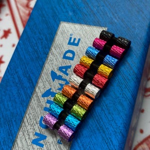 May include: A collection of colorful metal objects, each with a threaded end and a black band. The objects are in various colors, including black, pink, blue, yellow, red, silver, orange, green, and purple. The objects are arranged on a blue surface with the word "NOMAD" visible.