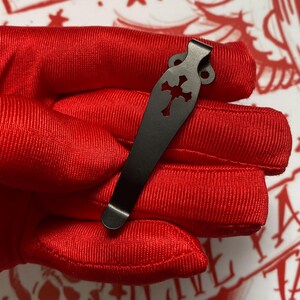 May include: A black metal pocket clip with a decorative cross cutout. The clip is designed to attach to a pocket or other item. The clip is shown against a red background.