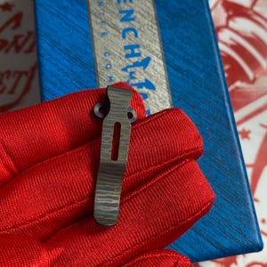 May include: A small, patterned metal clip with a rectangular slot and two circular holes. The clip is held by a red glove, with a blue and white box in the background. The word "Benchmade" is partially visible.