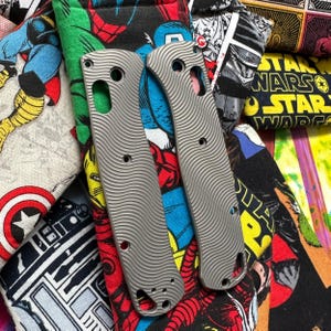 May include: Two silver-colored knife scales with a wavy, textured pattern. The scales have holes and cutouts for screws and a lanyard. They rest on a colorful fabric with comic book and Star Wars designs.